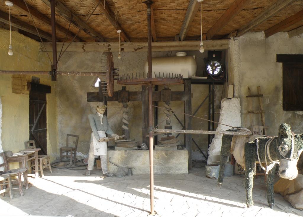 telpas stūrī stāvošais vīrieša manekens uzsāk riņķa danci ap olīvu spiedi, bet ēzeļa figūra - vilkt milzīgu dzirnu ratu. Redzams sens olīvu pārstrādes process. Foto: Iveta Krūmiņa